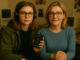 photograph features two versions of the same woman—one as a 90s teenager in headphones holding a Sony Walkman, the other as an adult in a blue jumper—seated together in a warmly lit, nostalgic study filled with vintage computers and old magazines.